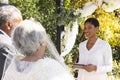 Biracial female marriage officiant using tablet with senior couple during wedding in sunny garden Royalty Free Stock Photo