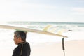 Biracial senior man carrying surfboard over head at beach against sky during sunny day