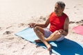 Biracial senior man with eyes closed meditating on yoga mat at sunny beach