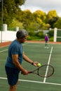 Biracial senior man serving tennis ball to senior wife while playing tennis in court against trees Royalty Free Stock Photo