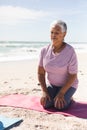 Biracial senior woman with eyes closed meditating while kneeling on yoga mat at sunny beach Royalty Free Stock Photo