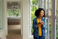 Biracial woman holding cup of coffee and looking out window at home Royalty Free Stock Photo