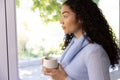 Biracial woman holding cup of coffee and looking out window at sunny home Royalty Free Stock Photo