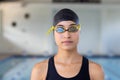Biracial young female swimmer standing indoors in front of a pool, wearing goggles Royalty Free Stock Photo