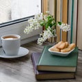 Bird cherry tree branches flowers, white cup of tea, cookies on close books on dark wooden background with row of books Royalty Free Stock Photo