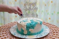 Birthday cake decorated with white and blue icing. Woman hand putting an ornament star. Floral curtain background. Front view