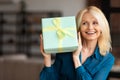 Birthday present. Joyful mature woman holding wrapped gift box near face guessing what inside, sitting on sofa at home