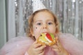 Birthday. Pretty, festively dressed red-haired baby in party hat enjoys delicious cake for her birthday, sitting at Royalty Free Stock Photo