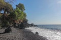 Black beach with lava pebbles and larger stone. Rock cliff of hardened volcanic lava. Slightly wavy sea, foamy water in the surf. Royalty Free Stock Photo