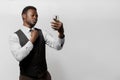 Black bearded african is looking to self through the camera on smartphone and straightens a tie. Man in white shirt and Royalty Free Stock Photo