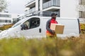 Black blue-collar worker - package delivery person - in front of white truck with cardboard package. Clothing - red Royalty Free Stock Photo