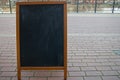 Black board with empty backdrop with traces of chalk in light brown wooden frame, standing on embankment on background of sidewalk Royalty Free Stock Photo