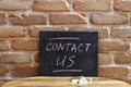 Black board with the phrase CONTACT US drown by hand on wooden table on brick wall background