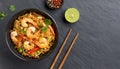 In a black bowl, noodles, veggies, and prawns are stir-fried. slate background. higher perspective