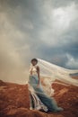 Black bride in waving long wedding dress and bridal veil stands on background of beautiful landscape. Royalty Free Stock Photo