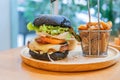 Black burger with meat patty, cheese, tomatoes, mayonnaise, french fries in a paper cup and glass of cold cola soda with ice.