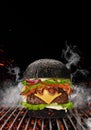 Black burger roasted on barbecue grill with flaming fire, sparks and smoke against black background. Beef cutlet, ham Royalty Free Stock Photo