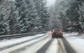 Black car drives on snow covered forest road during snowstorm, trees on both sides, view from car behind. Dangerous driving