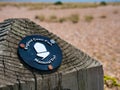 A black circular sign with white print and acorn logo affixed to a wooden post marks the way of the England Coast Path