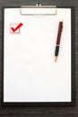 Black clipboard with white sheet of paper and metal pen isolated on dark wooden table. Checkbox with a tick on the page