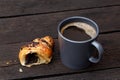 Black coffee in a blue-grey ceramic mug next to a half of chocolate croissant isolated on rustic dark brown wood table