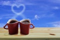 Black coffee in two pink cups heart shape with smoke is a heart shape on wooden floor and blue sky background