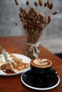 Black cup of hot cappuccino coffee with latte art on top on the wooden table with dessert Royalty Free Stock Photo