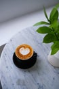 Black cup of tasty cappuccino with heart shaped latte art with vase with green tropical leaves on white table.