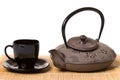 Black cup of tea on saucer and iron teapot on wooden mat.