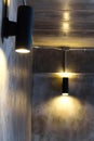 A black cylindrical wall lamp that shines up and down, splashes light onto the bare cement wall of the loft style house