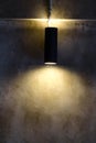 A black cylindrical wall lamp that shines up and down, splashes light onto the bare cement wall of the loft style house