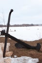 A black dark ship anchor is on the ground leaning on rocks against wide snowy empty landscape with far away trees in the fog and Royalty Free Stock Photo