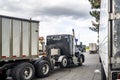 Black day cab big rig semi truck tractor with bulk semi trailer moving to truck stop parking lot exit with line of another semi Royalty Free Stock Photo