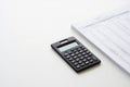 A black electronic calculator with a small screen sits on a clean white background next to a stack of papers isolated on white Royalty Free Stock Photo