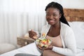 Black Expectant Woman Eating Fresh Vegetable Salad, Sitting On Bed At Home Royalty Free Stock Photo