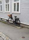 A black fashionable bicycle with a basket on the handlebars stands against the wall of an old wooden white house