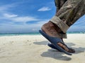 The black feet of an old man wearing blue sandals and military pants walking along the white sandy beach.