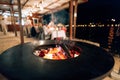 Black flat grill, round shape, with a hole with a fire in the center. In the background of the people dining at the