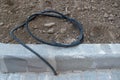 Black flexible corrugated pipe coiled lying on gray soil at curb, other end goes underground, intended for cable pulling