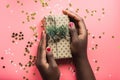 Black girl is holding a gift of Kraft paper on a pink background with confetti. Womans hands holding gift or present box decorated Royalty Free Stock Photo