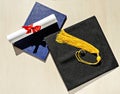 Black graduate cap with yellow tassel, blue diploma and paper scroll tied with red ribbon with bow on beige wooden background