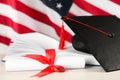 Black graduation hat, diploma and book on white wooden table against American flag Royalty Free Stock Photo