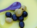 Olives on a yellow plate with a lilac spoon.