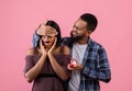 Black guy covering his sweetheart`s eyes, giving her engagement ring, making marriage proposal on pink background Royalty Free Stock Photo