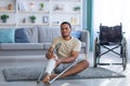 Black guy sitting on floor after falling with crutches, suffering from pain in broken leg, wheelchair standing nearby Royalty Free Stock Photo