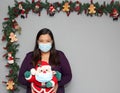 Black hair latin woman with protection mask and santaclaus doll in christmas decoration, new normal covid-19