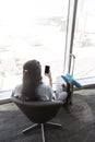 Black-haired Caucasian woman sits in an airport lounge seat, checking her cell phone. She also has her carry on