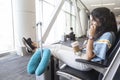 Black-haired Caucasian woman is sitting in an airport lounge seat with her passport and a coffee in her hands. She also has her