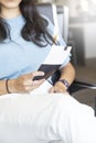 Black-haired Caucasian woman is sitting in an airport lounge seat with her passport in her hands. She also has her carry on