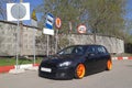 Black hatchback with orange wide wheels tuned by air suspencion parked under different road signs